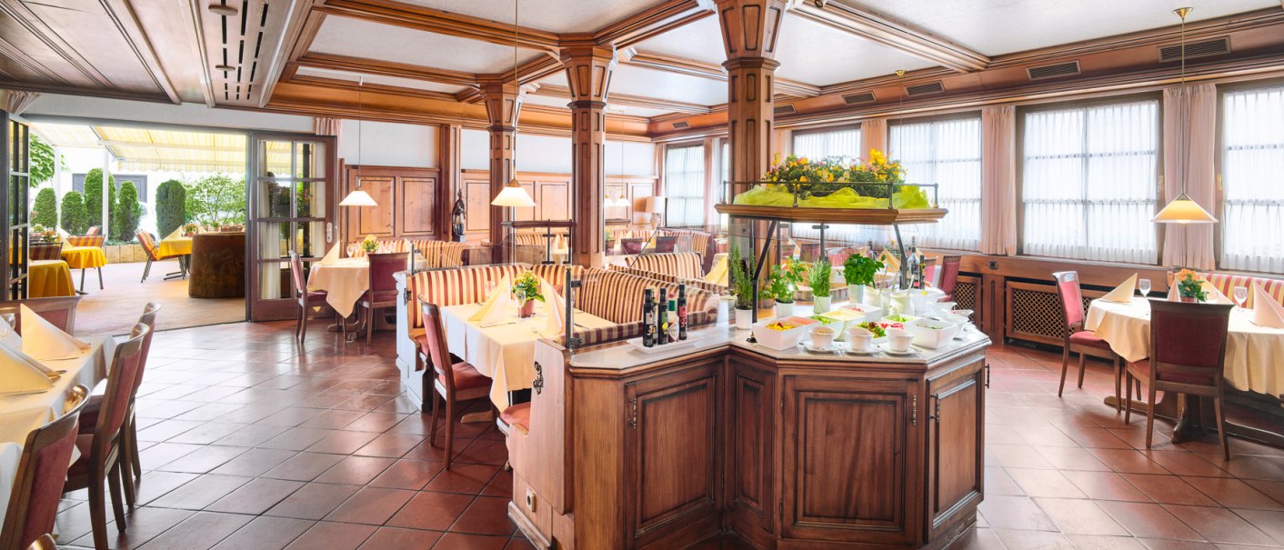 Elegantes Restaurant mit Holzmöbeln, gedeckten Tischen und einem Buffet. Große Fenster und eine Terrasse im Hintergrund sorgen für viel Licht., © Hirsch Hotel