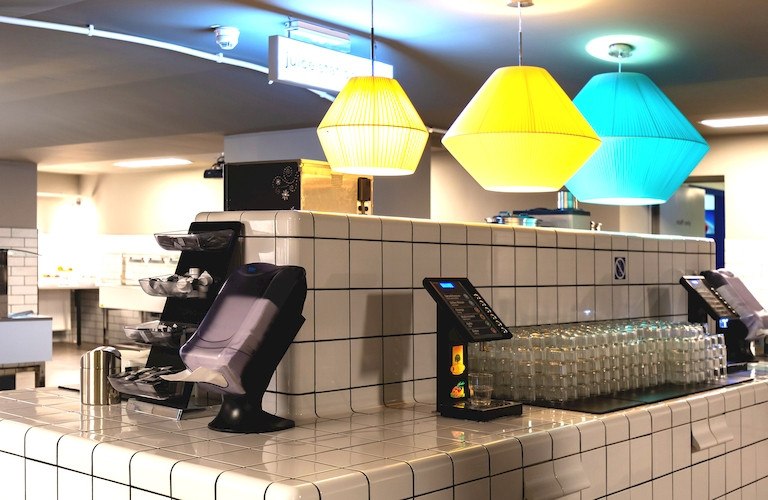 Modern breakfast bar with colorful lamps, stacked glasses and a juice dispenser. Tile design and subtle lighting., © A&O Hostels Marketing GmbH Modern breakfast bar with colorful lamps, stacked glasses and a juice dispenser. Tile design and subtle lighting., © A&O Hostels Marketing GmbH