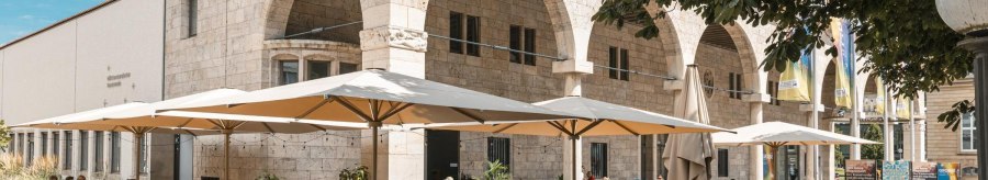 A café with large parasols and guests sitting at tables outside. The building has arches and is made of stone., © Stuttgart Marketing GmbH, Sarah Schmid