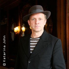 Ein Mann mit Hut und gestreiftem Shirt steht in einem eleganten, dunklen Raum., © links im Bild Ein Mann mit Hut und gestreiftem Shirt steht in einem eleganten, dunklen Raum., © links im Bild