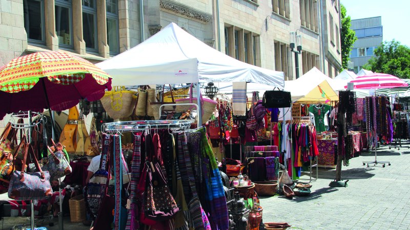 Ein lebhafter Markt mit bunten Ständen, die Kleidung, Taschen und Accessoires anbieten. Weiße und bunte Sonnenschirme spenden Schatten., © Forum der Kulturen Stuttgart e. V. Ein lebhafter Markt mit bunten Ständen, die Kleidung, Taschen und Accessoires anbieten. Weiße und bunte Sonnenschirme spenden Schatten., © Forum der Kulturen Stuttgart e. V.