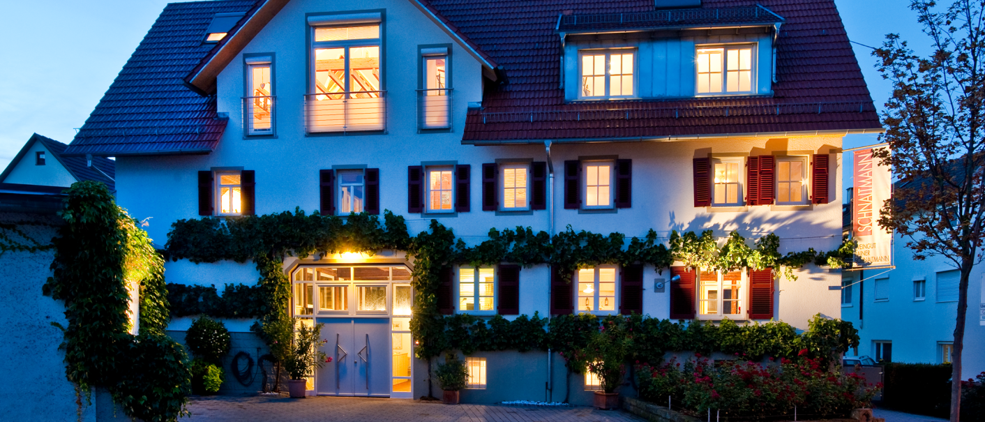 Das Weingut Schnaitmann bei Dämmerung, ein beleuchtetes Gebäude mit roten Fensterläden und Efeu an der Fassade. Ein Schild mit dem Namen hängt rechts., © Weingut Schnaitmann