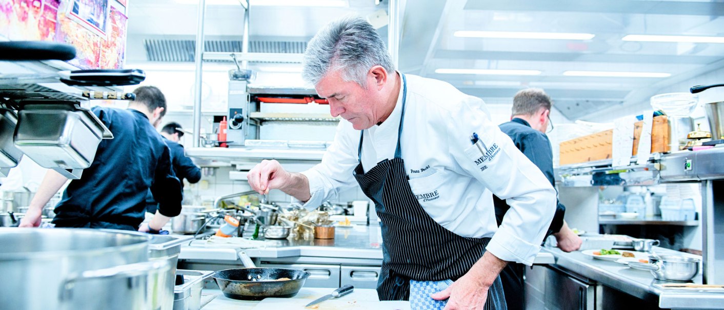 Ein Koch in einer modernen Küche würzt ein Gericht. Im Hintergrund arbeiten weitere Köche. Die Küche ist gut ausgestattet und sauber., © Landhaus Franz Feckl Ein Koch in einer modernen Küche würzt ein Gericht. Im Hintergrund arbeiten weitere Köche. Die Küche ist gut ausgestattet und sauber., © Landhaus Franz Feckl