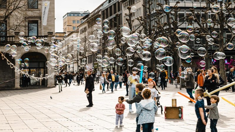 Ein Stra&szlig;enk&uuml;nstler erzeugt Seifenblasen auf einer belebten Stra&szlig;e. Kinder spielen begeistert mit den Blasen, w&auml;hrend Passanten zuschauen., &copy; Stuttgart-Marketing GmbH, Sarah Schmid