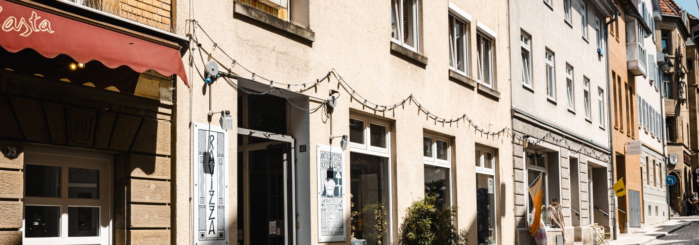 Eine sonnige Gasse mit Kopfsteinpflaster, ges&auml;umt von Gesch&auml;ften wie einem Antiquariat. Lichterketten h&auml;ngen &uuml;ber den Eing&auml;ngen., &copy; SMG Stuttgart Marketing GmbH - Sarah Schmid
