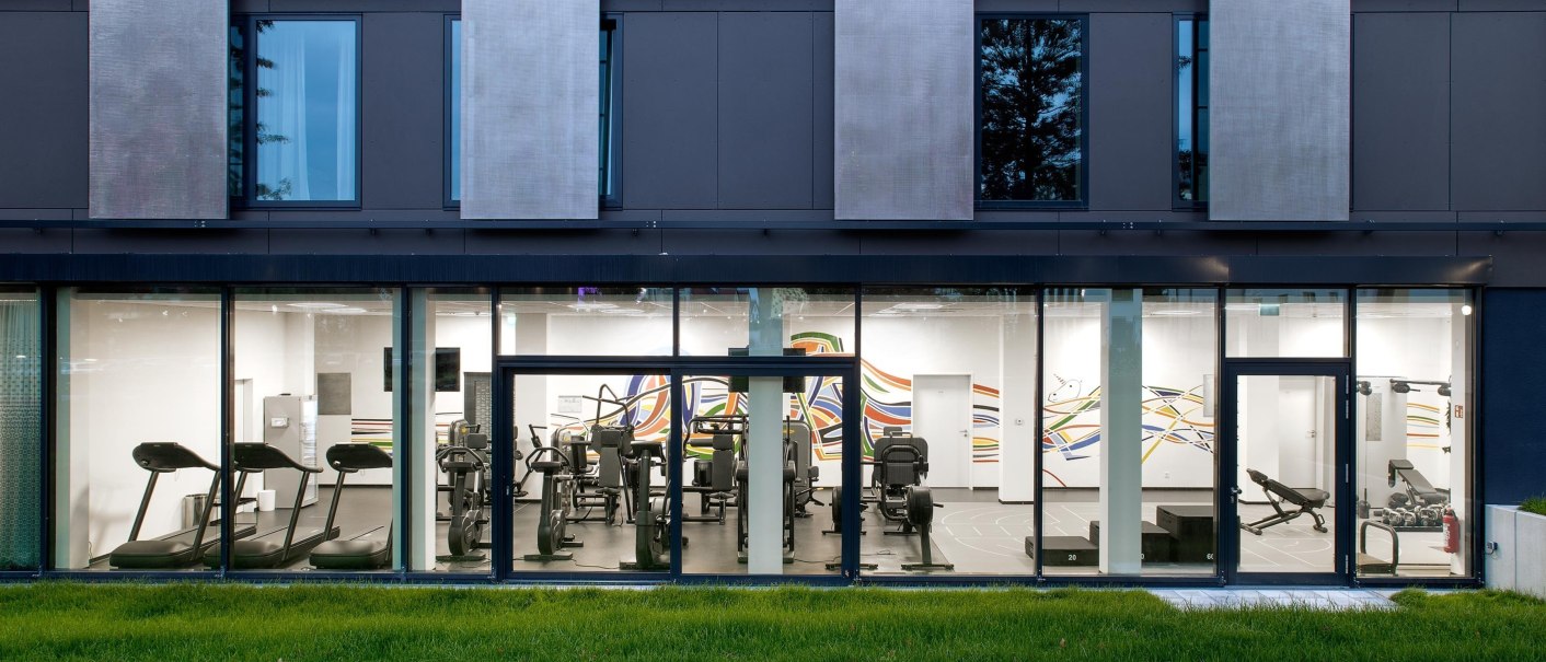 Modern gym façade with large windows offering a view of the treadmills and fitness equipment inside. Green meadow in the foreground., © Hotel am Remspark Modern gym façade with large windows offering a view of the treadmills and fitness equipment inside. Green meadow in the foreground., © Hotel am Remspark