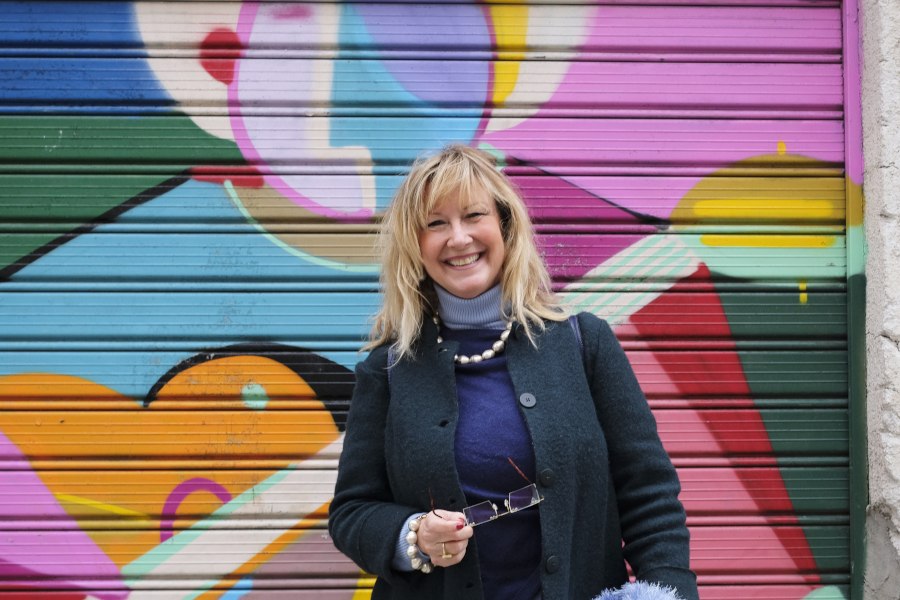 Eine lächelnde Frau steht vor einer bunten Graffiti-Wand. Sie trägt einen dunklen Mantel und hält eine Brille in der Hand., © Foto: privat Eine lächelnde Frau steht vor einer bunten Graffiti-Wand. Sie trägt einen dunklen Mantel und hält eine Brille in der Hand., © Foto: privat