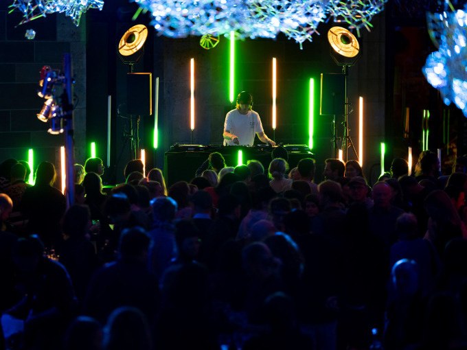 A DJ plays at a party, surrounded by colorful lights and a dancing crowd. The atmosphere is lively and full of energy., &copy; Landesmuseum W&uuml;rttemberg