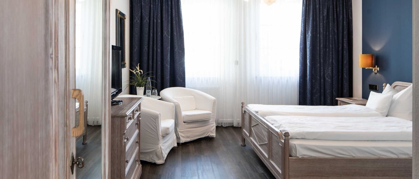An elegant hotel room with a double bed, white armchairs, dark curtains, and wooden furniture. Bright lighting and stylish decor., &copy; Flairhotel Vier Jahreszeiten