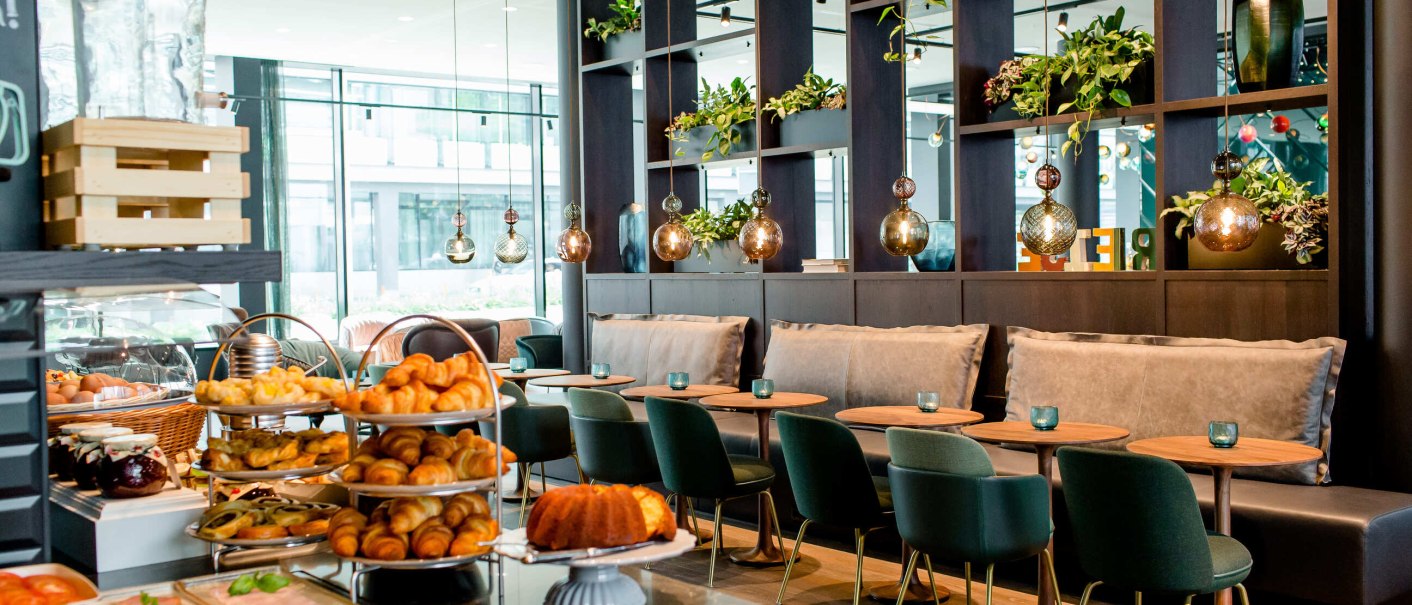 Elegantes Frühstücksbuffet mit Croissants, Kuchen und Gemüse in einem stilvollen Restaurant mit grünen Stühlen und hängenden Lampen., © Motel One - Stuttgart Hauptbahnhof