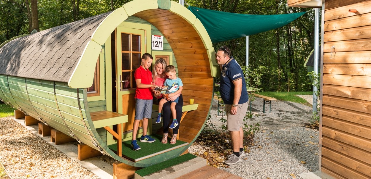 Eine Familie sitzt vor einem fassförmigen Holzhaus im Freien. Ein Mann steht daneben. Umgeben von Bäumen und einem Holzweg., © Remsfässer