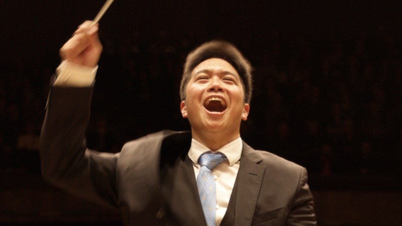 Ein l&auml;chelnder Dirigent in einem Anzug dirigiert mit einem Taktstock vor einem dunklen Hintergrund., &copy; Jochen Stall