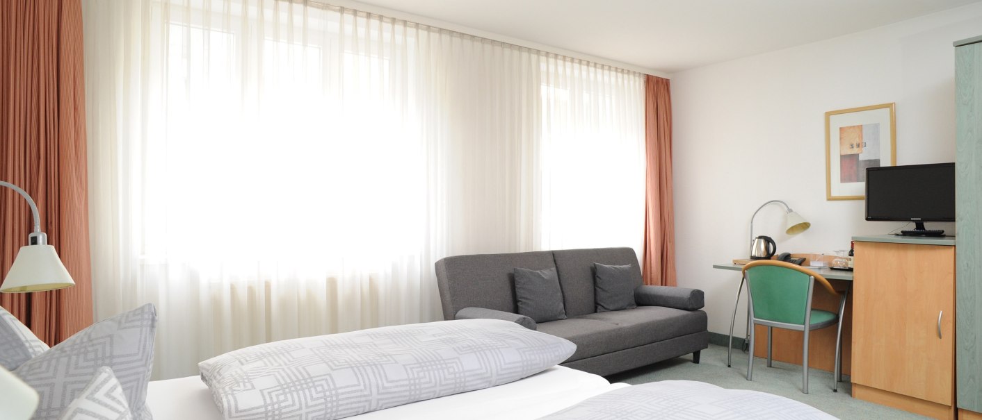 Bright hotel room with double bed, gray sofa, desk with lamp and TV. Large windows with curtains let in plenty of light., © TOMAS