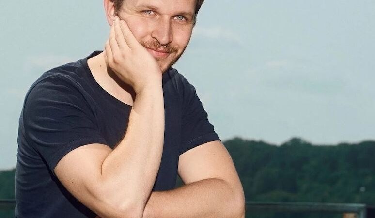 Ein Mann in einem schwarzen T-Shirt lehnt sich lächelnd auf ein Geländer. Im Hintergrund sind Bäume und ein klarer Himmel zu sehen., © Rosenau Kultur e.V. Ein Mann in einem schwarzen T-Shirt lehnt sich lächelnd auf ein Geländer. Im Hintergrund sind Bäume und ein klarer Himmel zu sehen., © Rosenau Kultur e.V.