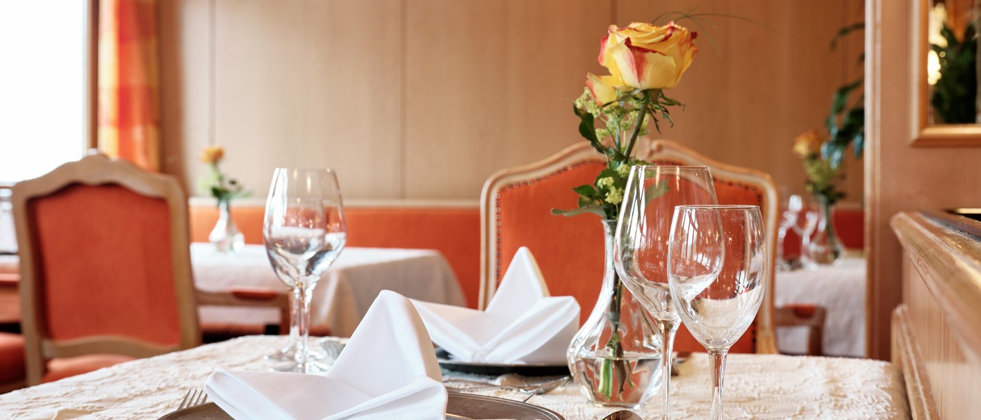 Eleganter Tisch im Restaurant mit weißer Tischdecke, Weingläsern, Besteck und gelber Rose in Vase. Holzwände und orangefarbene Stühle im Hintergrund., © Badhotel Restaurant Stauferland