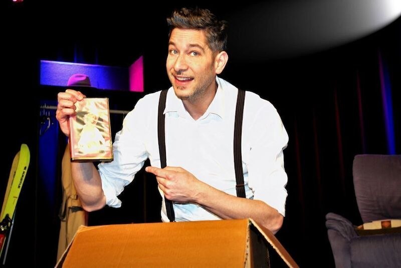 Ein Mann auf einer B&uuml;hne h&auml;lt eine DVD-H&uuml;lle und zeigt darauf. Er tr&auml;gt ein wei&szlig;es Hemd mit Hosentr&auml;gern und l&auml;chelt in die Kamera., &copy; Theaterhaus Stuttgart e.V.