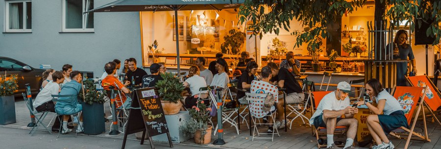 Abendliche Szene auf einer Terrasse mit vielen Menschen, die unter einem Sonnenschirm sitzen und sich unterhalten. Im Hintergrund leuchtet ein Caf&eacute;., &copy; Radcaf&eacute; Fietsen