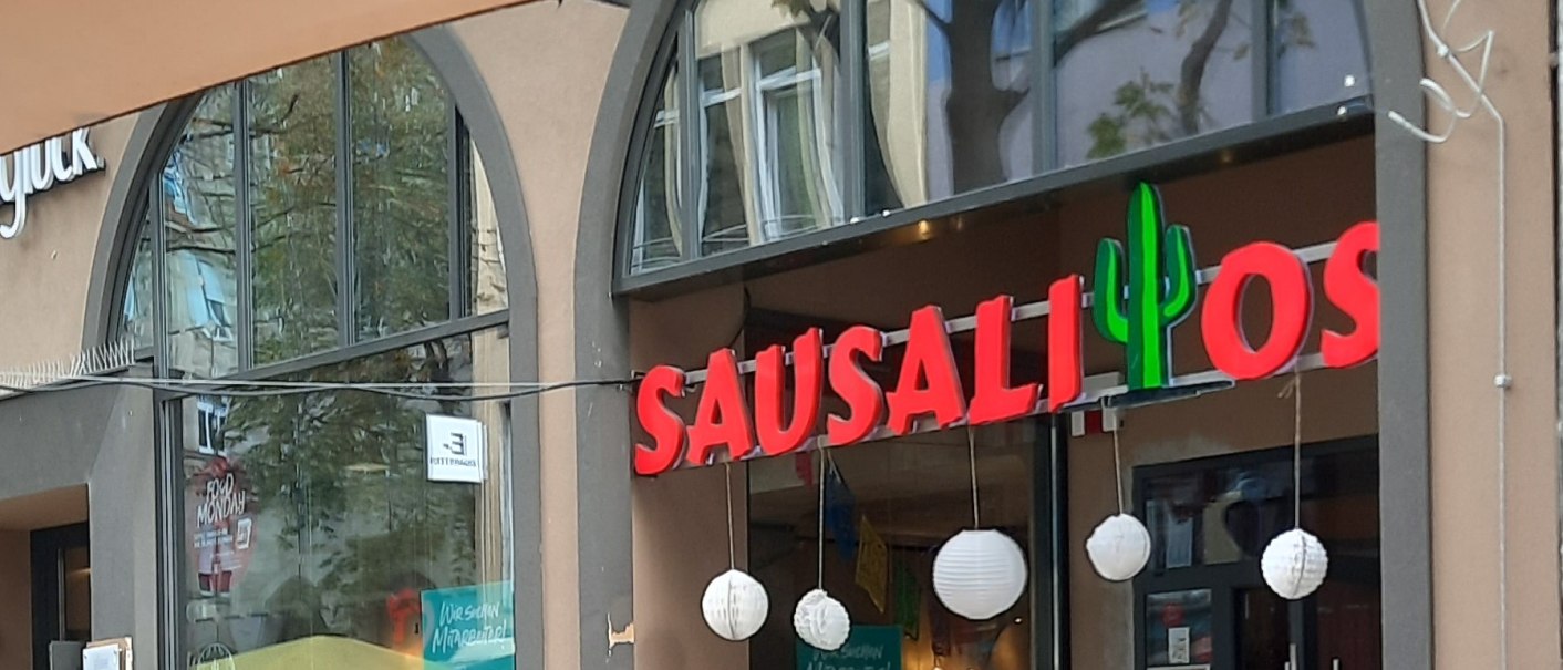 Entrance to the Sausalitos restaurant with red lettering and cactus logo. A sign advertises Taco Tuesday. Outdoor area with wooden tables., &copy; SMG
