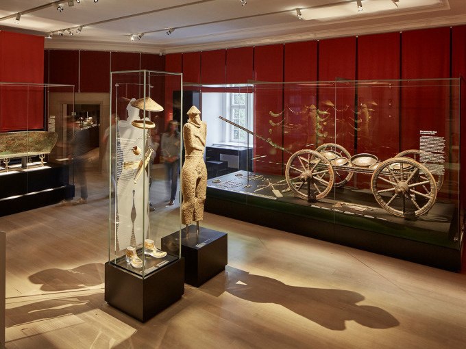 Museumsausstellung mit antiken Artefakten, darunter eine Statue und ein Streitwagen in einem Raum mit roten Wänden., © Landesmuseum Württemberg Museumsausstellung mit antiken Artefakten, darunter eine Statue und ein Streitwagen in einem Raum mit roten Wänden., © Landesmuseum Württemberg