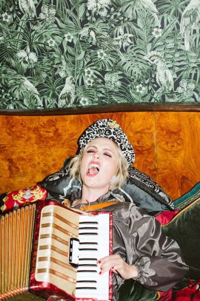 A person in a leopard hat plays an accordion and sings passionately in front of a tropical wallpaper., &copy; Rosenau Kultur e.V.