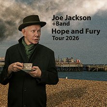 Ein Mann mit Hut und Schal h&auml;lt eine Tasse vor einem Strand mit einem Pier im Hintergrund. Text: Joe Jackson + Band, Hope and Fury Tour 2026., &copy; links im Bild