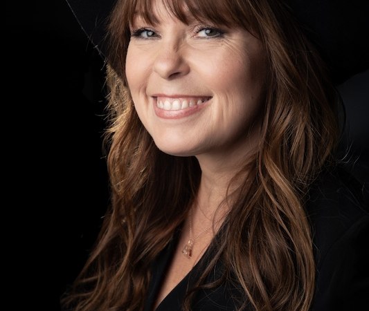A woman with long brown hair and a black hat smiles at the camera against a dark background., &copy; Rosenau Kultur e.V.