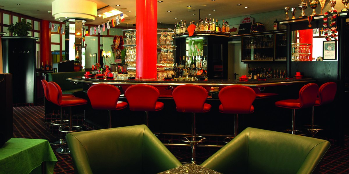 Elegante Bar mit roten Hockern, zentraler Theke und dekorativen Flaschen. Internationale Flaggen und Trophäen schmücken den Raum., © © H-Hotels.com Elegante Bar mit roten Hockern, zentraler Theke und dekorativen Flaschen. Internationale Flaggen und Trophäen schmücken den Raum., © © H-Hotels.com