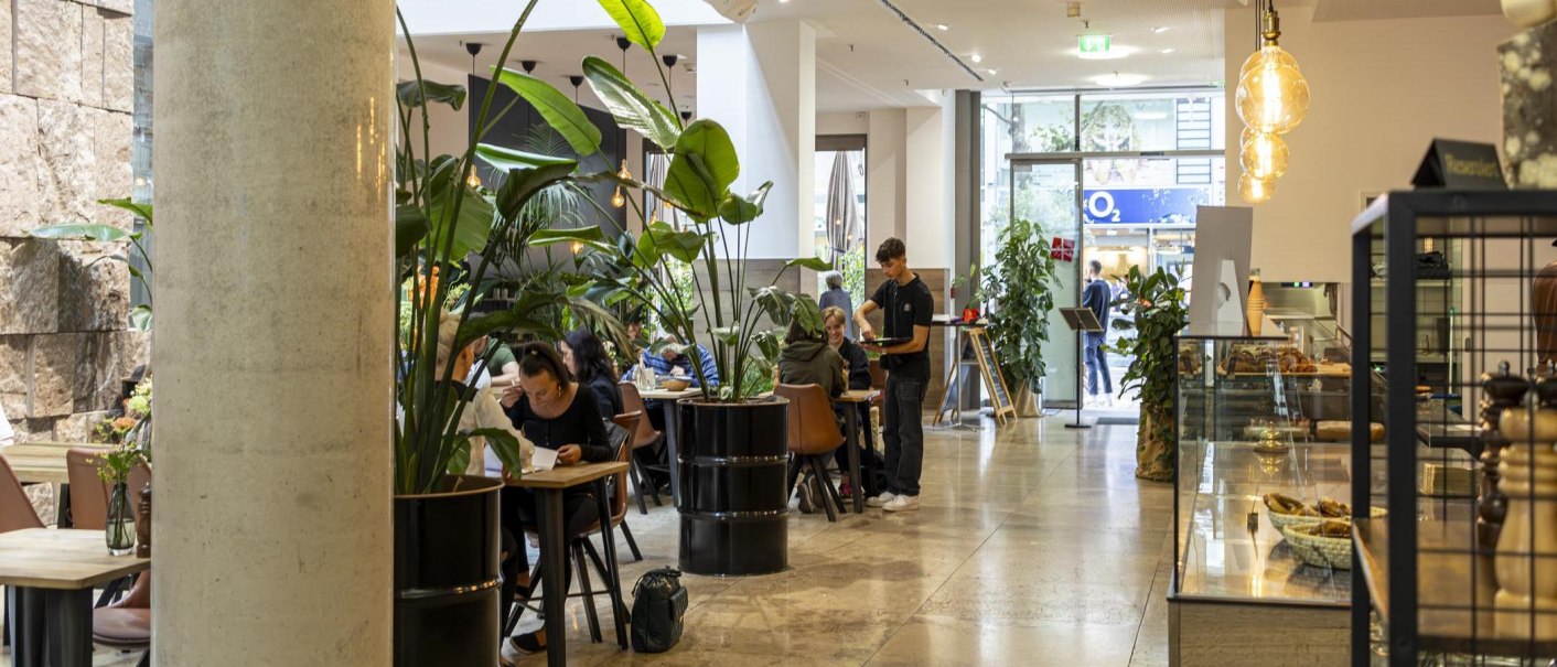 Modernes Café mit großen Pflanzen, Tischen und einem Bediener. Menschen sitzen und genießen ihre Zeit. Helle, einladende Atmosphäre., © SMG, Sarah Schmid Modernes Café mit großen Pflanzen, Tischen und einem Bediener. Menschen sitzen und genießen ihre Zeit. Helle, einladende Atmosphäre., © SMG, Sarah Schmid