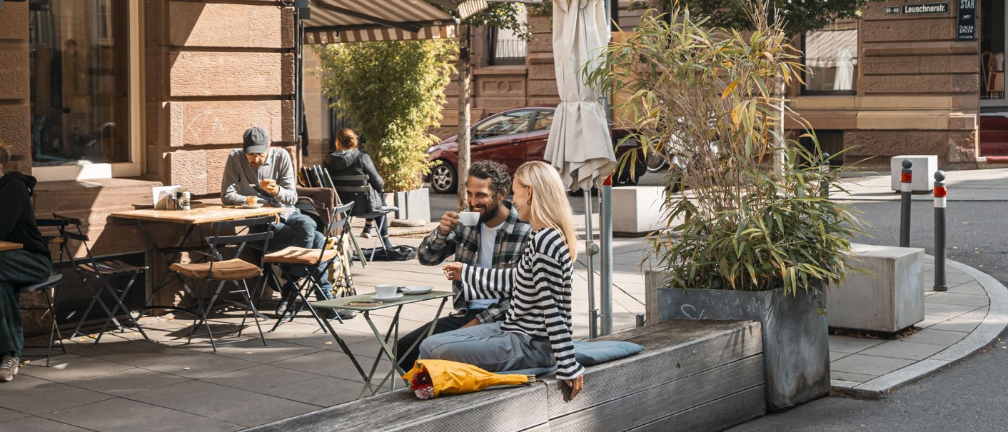 People enjoying coffee in front of a café in the sun. A couple is sitting on a bench, others at tables. Plants and parasols decorate the scene., © SMG, Sarah Schmid