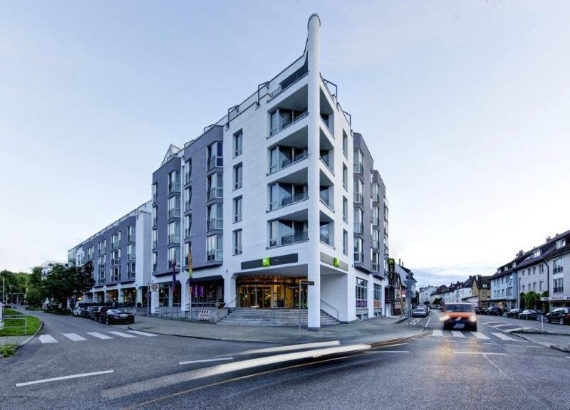 Modernes Hotelgebäude an einer Straßenecke mit Glasfassade und Balkonen, beleuchtet bei Dämmerung. Vorbeifahrende Autos erzeugen Lichtspuren., © Ibis Styles Stuttgart Modernes Hotelgebäude an einer Straßenecke mit Glasfassade und Balkonen, beleuchtet bei Dämmerung. Vorbeifahrende Autos erzeugen Lichtspuren., © Ibis Styles Stuttgart