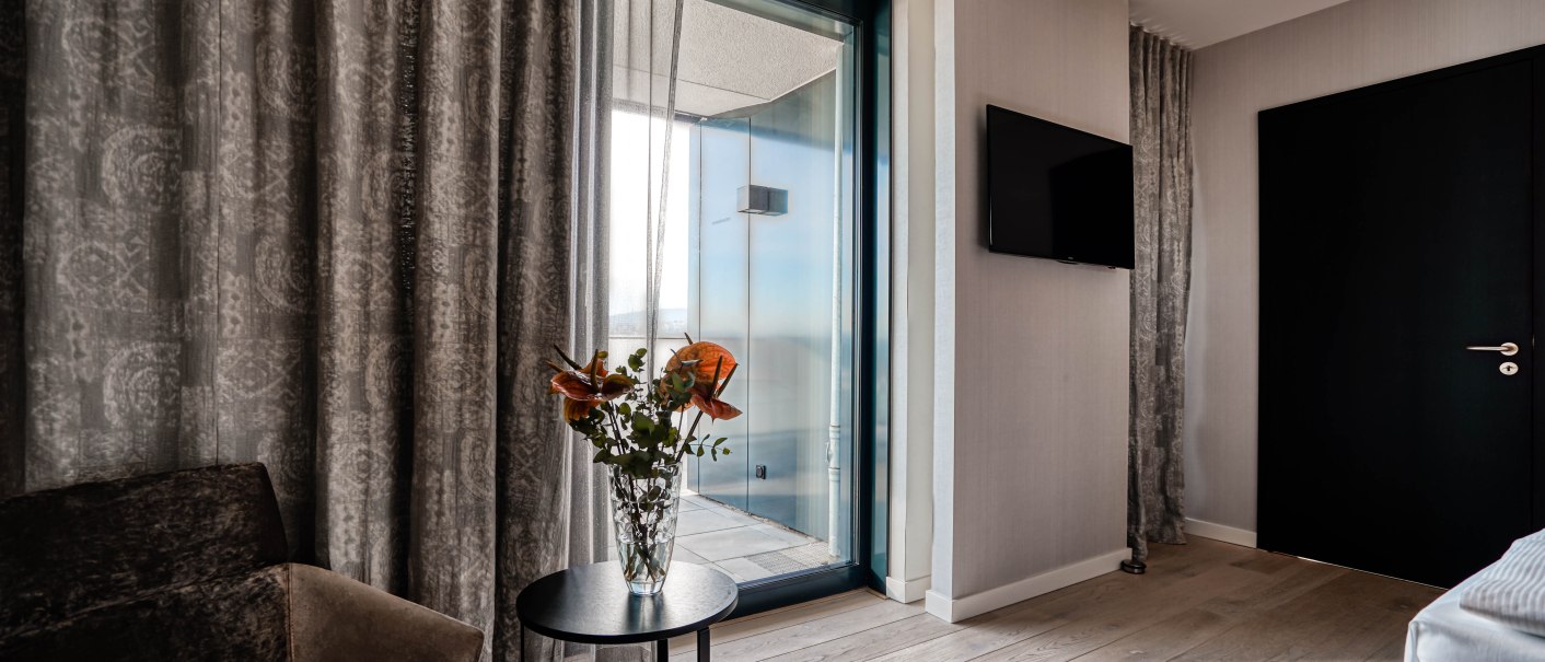 Elegantes Hotelzimmer mit Balkon, Vorhängen, Sessel, Tisch mit Blumen und Flachbildfernseher an der Wand., © CLOUD N°7 APARTMENTS GmbH