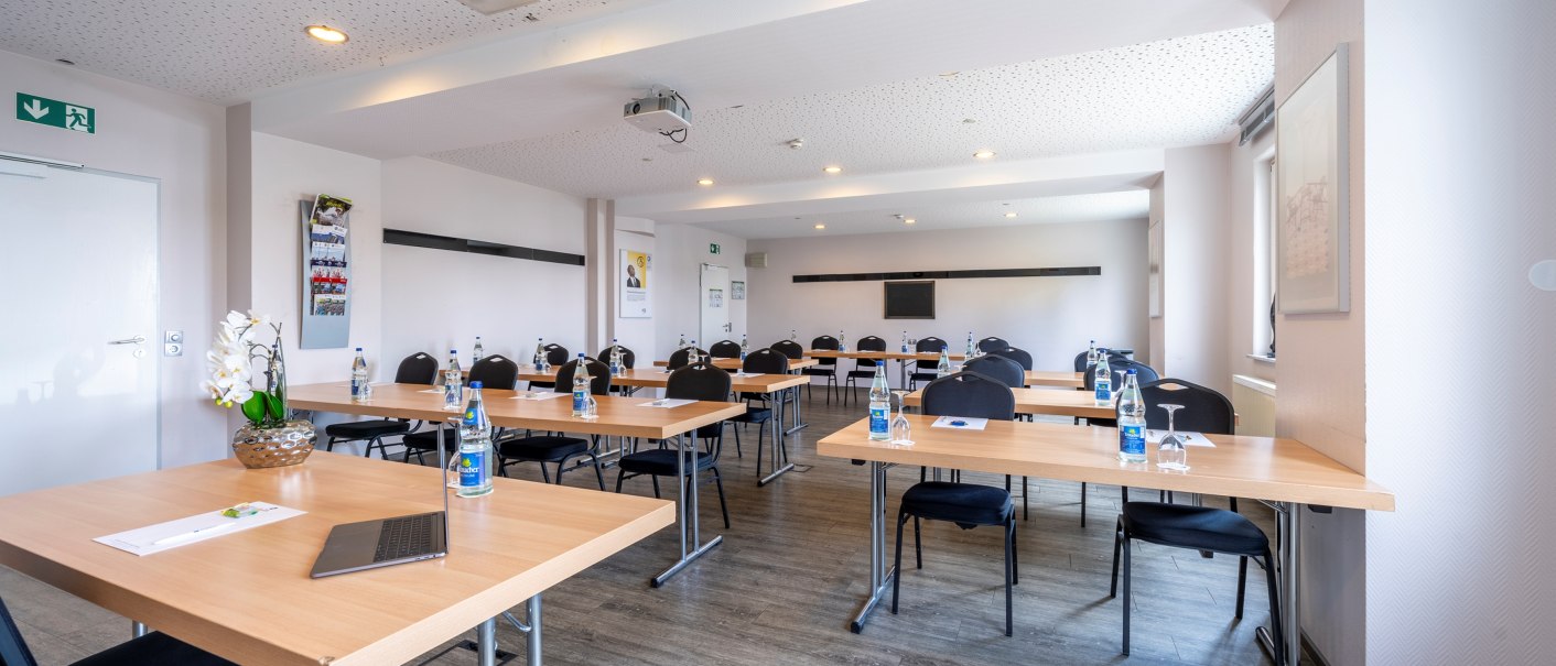 A bright meeting room with tables, chairs and bottles of water. A projector hangs from the ceiling and there are posters and an emergency exit sign on the walls., &copy; Plaza INN Stuttgart-Filderstadt