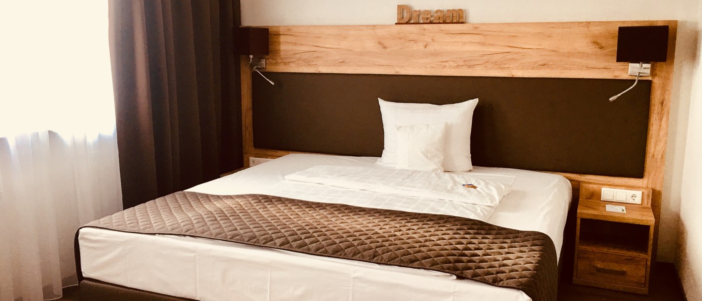 Modern hotel room with double bed, brown curtains and wooden furniture. 'Dream' is written on the headboard. Bright, cozy atmosphere., © TOMAS Modern hotel room with double bed, brown curtains and wooden furniture. 'Dream' is written on the headboard. Bright, cozy atmosphere., © TOMAS