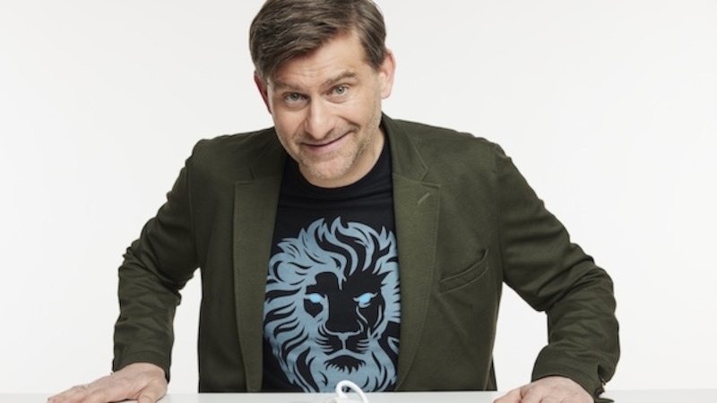 Mann in olivgr&uuml;nem Sakko mit L&ouml;wen-T-Shirt lehnt sich &uuml;ber Tisch, l&auml;chelt in die Kamera., &copy; Renitenztheater Stuttgart e.V.