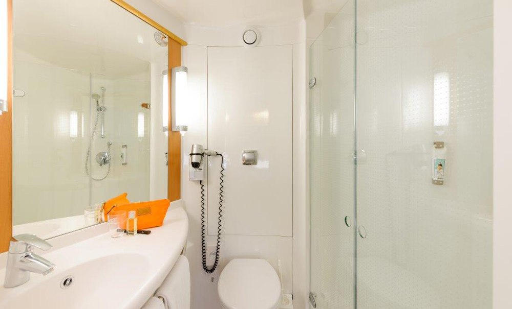 Modernes Badezimmer mit Glasdusche, Waschbecken, großem Spiegel und Toilette. Ein Föhn hängt an der Wand, und eine orangefarbene Tasche steht auf dem Waschbecken., © Ibis Stuttgart Airport Messe