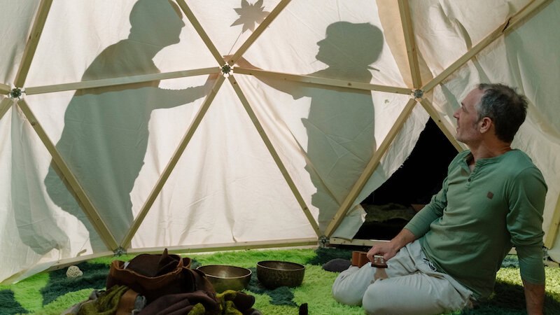 Ein Mann sitzt in einem Zelt auf grünem Boden, während zwei Schatten an der Zeltwand zu sehen sind. Im Zelt liegen Klangschalen., © FITZ Das Theater animierter Formen