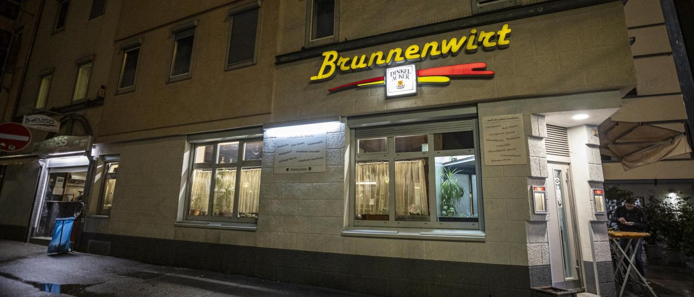 Das Restaurant Brunnenwirt bei Nacht, beleuchtet mit gelbem Schriftzug. Ein Mann steht an einem Tisch vor dem Eingang., &copy; SMG, Sarah Schmid