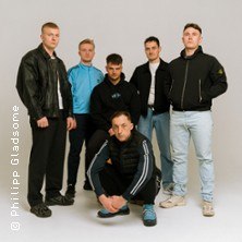 Sechs Männer in sportlicher und lässiger Kleidung posieren vor einem neutralen Hintergrund. Einer hockt vorne, die anderen stehen dahinter., © links im Bild Sechs Männer in sportlicher und lässiger Kleidung posieren vor einem neutralen Hintergrund. Einer hockt vorne, die anderen stehen dahinter., © links im Bild