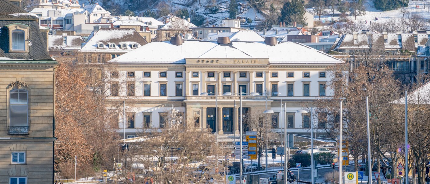 The StadtPalais in Stuttgart can be seen in winter, with a snow-covered roof and surrounding buildings. Trees without leaves in the foreground., © SMG Thomas Niedermüller