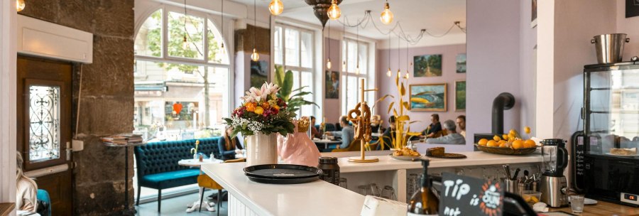 Gem&uuml;tliches Caf&eacute; mit Blumen und Brezeln auf der Theke, blauen Sofas und Kunst an den W&auml;nden. Gro&szlig;e Fenster lassen viel Licht herein., &copy; SMG, Sarah Schmid
