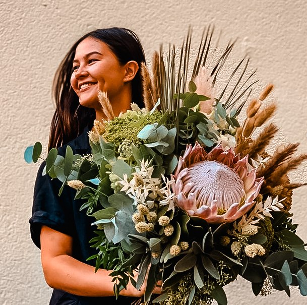Eine lächelnde Frau hält einen großen Blumenstrauß mit Protea und verschiedenen Gräsern vor einer beigen Wand., © Merz & Benzing GmbH