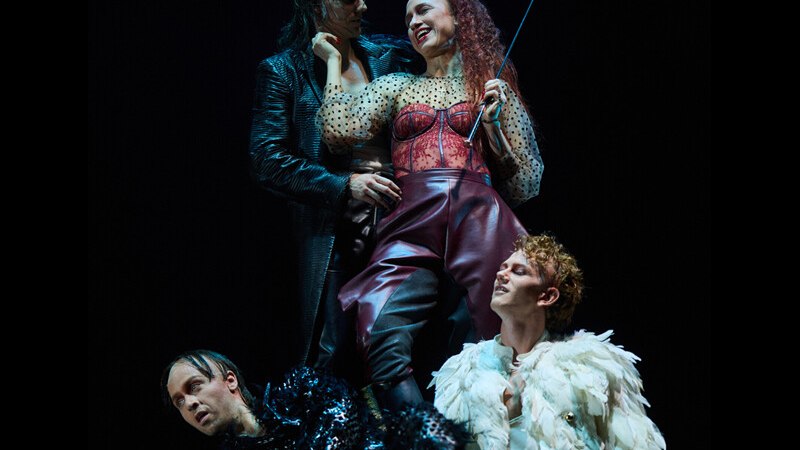 Vier Personen in extravaganten Kostümen posieren auf einer Bühne. Eine Frau in rotem Outfit hält einen Stock, umgeben von drei Männern in schwarzen und weißen Kostümen., © Württembergische Staatstheater Stuttgart Vier Personen in extravaganten Kostümen posieren auf einer Bühne. Eine Frau in rotem Outfit hält einen Stock, umgeben von drei Männern in schwarzen und weißen Kostümen., © Württembergische Staatstheater Stuttgart