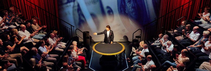 Ein Magier steht auf einer Bühne mit rotem Vorhang, umgeben von applaudierendem Publikum. Im Hintergrund sind vergrößerte Geldscheine projiziert., © STROTMANNS Magic Lounge