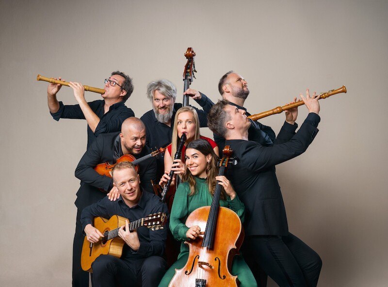 Eight musicians pose with various instruments, including flutes, guitar and cello, in front of a neutral background. They appear cheerful and creative., &copy; Theaterhaus Stuttgart e.V.