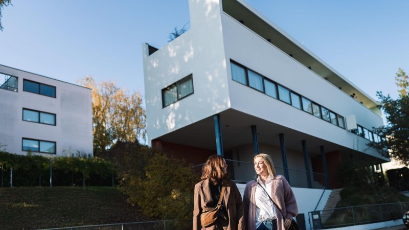 Zwei Frauen spazieren vor einem modernen, wei&szlig;en Geb&auml;ude der Wei&szlig;enhofsiedlung in Stuttgart. Der Himmel ist klar und blau., &copy; SMG, wpsteinheisser