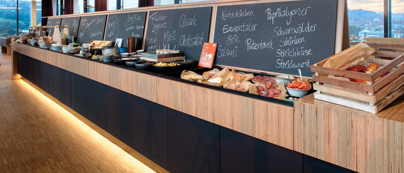 A rich breakfast buffet with various dishes and boards describing what is on offer. In the background are windows with a view., © Hotel am Remspark A rich breakfast buffet with various dishes and boards describing what is on offer. In the background are windows with a view., © Hotel am Remspark