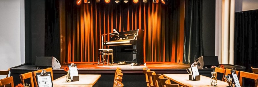Ein Veranstaltungsraum mit Bühne, Klavier und Mikrofon. Tische mit Blumen und Menükarten stehen bereit. Die Bühne ist in warmes Licht getaucht., © Rosenau e. V., Stuttgart Ein Veranstaltungsraum mit Bühne, Klavier und Mikrofon. Tische mit Blumen und Menükarten stehen bereit. Die Bühne ist in warmes Licht getaucht., © Rosenau e. V., Stuttgart