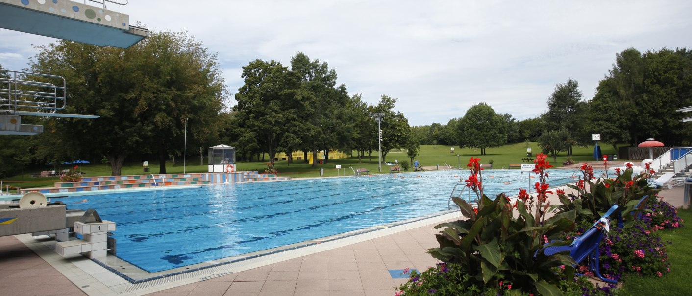 Ein Freibad mit einem großen Schwimmbecken, umgeben von grünen Bäumen und bunten Blumenbeeten. Der Himmel ist leicht bewölkt., © Stuttgarter Bäder
