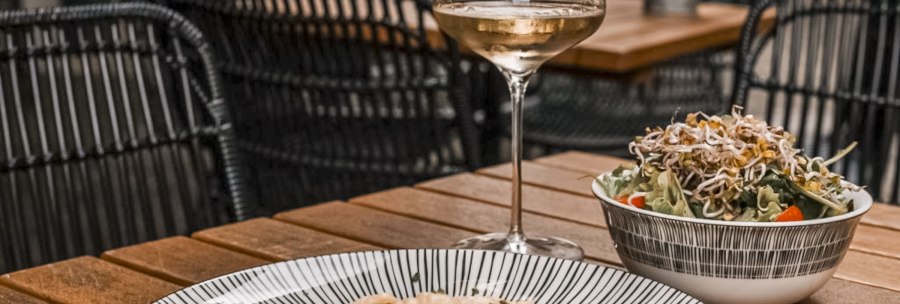 A glass of white wine, a plate of pasta and a salad on a wooden table in a restaurant with woven chairs., © SMG, itchyfeet-travel.de