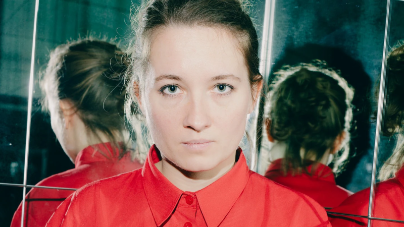 Eine Frau in einem roten Hemd steht vor einem Spiegel, der ihr Gesicht und ihren R&uuml;cken reflektiert. Der Hintergrund ist dunkel., &copy; Sophia Hegelwald